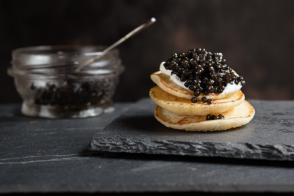 Black sturgeon caviar on small pancakes blinis with sour cream and a glass jar with caviar on a slate (image supplied)